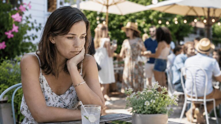 Frau sitzt nachdenklich allein an einem Tisch bei einer sommerlichen Gartenfeier, während im Hintergrund Menschen miteinander sprechen.